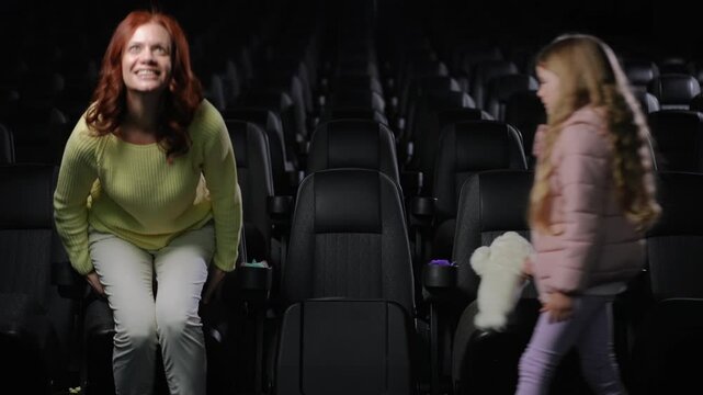 A family of three arrives at an empty cinema hall and settles into the first row. The mother, wearing a yellow sweater, smiles as she sits down, while the father, dressed in a green sweater, helps
