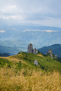 A Picturesque Scene Of Rolling Green Hills, Towering Rock Formations, And Golden Grasses, Set Against A Backdrop Of Layered Blue Mountains And A Cloudy Sky