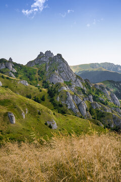 A Serene Mountain Landscape Featuring Rugged Rock Formations, Lush Green Hills, Golden Grasses In The Foreground, And A Clear Blue Sky With Wispy Clouds