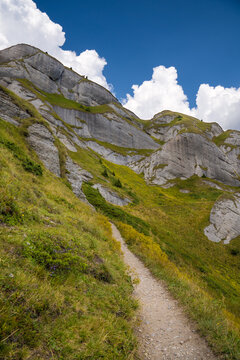 A Serene Mountain Path Winds Through Lush Greenery And Rugged Rock Formations Under A Vibrant Blue Sky Dotted With Puffy White Clouds