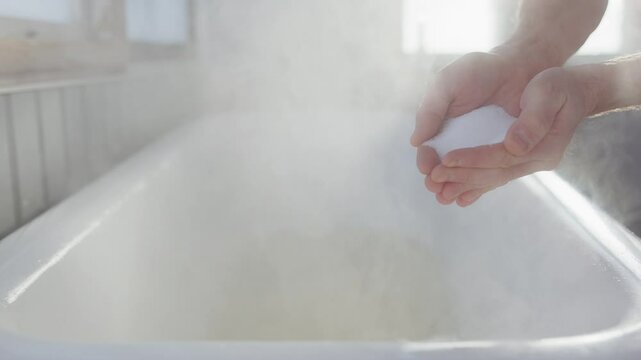 Cupped hands drop white epsom salts into steamy bath, telephoto