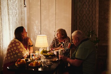Happy elderly couple and their adult daughter celebrate holiday at the table, family of three have dinner in lamplight at home in the kitchen