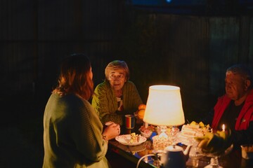 Happy elderly couple and their adult daughter celebrate holiday in the courtyard of the house, family of three have dinner in lamplight