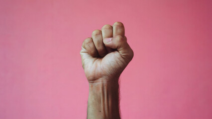 A human hand raised in a clenched fist against a pink background. The hand appears to be of an adult, with visible veins and a slightly weathered skin texture