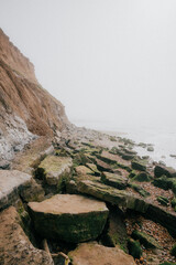 A rocky shoreline with a foggy sky in the background. The rocks are covered in moss and the water is choppy