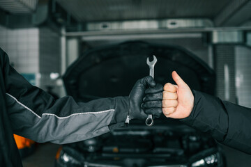 Close-up of a mechanic's gloved hand holding a wrench and a customer's hand giving a thumbs-up,...