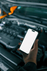 Mechanic holding a smartphone with a blank white screen in front of an open car engine for diagnostics or maintenance. Indoor workshop setting