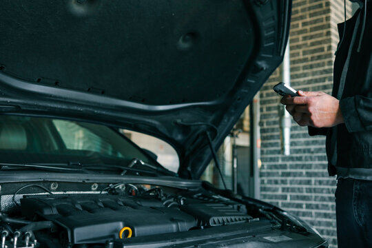 Mechanic using a smartphone while inspecting an engine under the open hood of a car in a professional garage. Modern technology in automotive diagnostics and repair