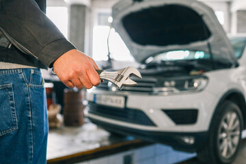Mechanic holding an adjustable wrench in a workshop. A white car with its hood open is visible in the background, indicating repair or maintenance work