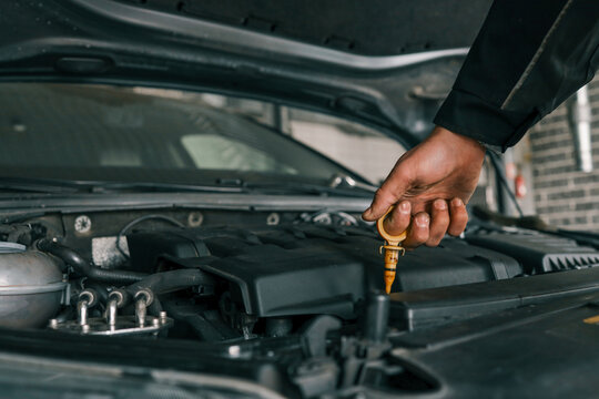 Close-up of a mechanic's hand holding an oil dipstick, checking engine oil levels in a vehicle's open hood. Represents routine car maintenance and servicing