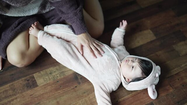 In a warm winter Japanese apartment room, a 6-month-old Japanese girl wearing fluffy clothes and her mother are sitting on the floor holding their sleeping baby. Slow motion video.
