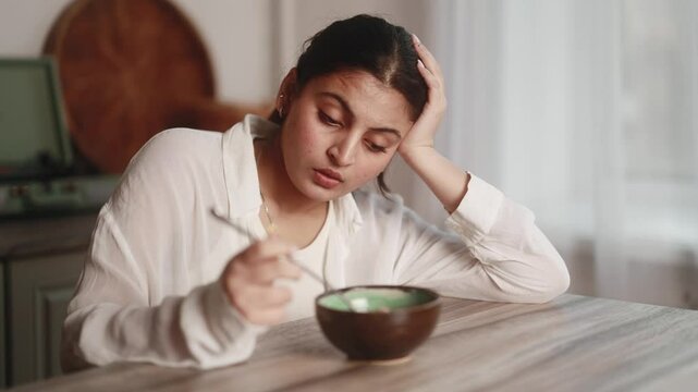 Portrait of worried young indian woman no appetite due to personal or work problems sitting at table on the home kitchen Dissatisfied female looks at the not tasty breakfast indoors Bad morning