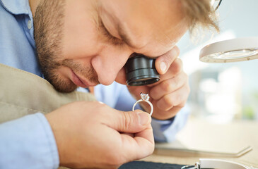 Closeup portrait of young male jewellery appraiser inspecting gold ring with diamond looking through magnifying glass working at workshop. Male jeweler and goldsmith evaluating jewelry.