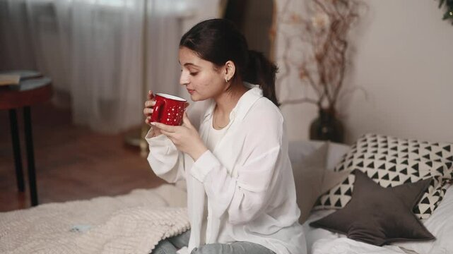 Portrait of charming young indian woman sitting on bed take sip drinking hot tea or coffee at cozy home Calm relaxed female enjoying weekend leisure time alone in the bedroom indoors