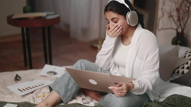 Boring unmotivated young indian woman student listening online lecture by laptop computer sitting on bed at home Tired girl with uninteresting lesson on remote distance education indoors