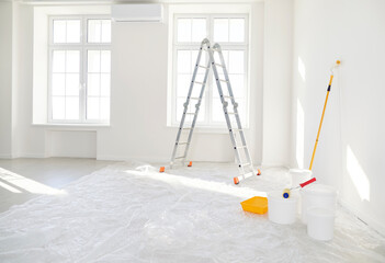 Step ladder, buckets with paint roller standing in empty light room in new house or flat with freshly painted white walls. Renovation process in apartment. Home repair and moving day concept.