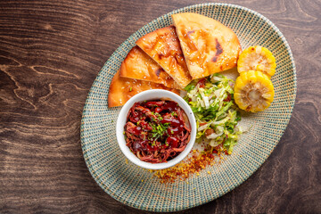 A vibrant plate featuring crispy flatbreads, tender pulled meat in sauce, fresh salad, and grilled corn, all beautifully arranged on a textured platter against a wooden background.
