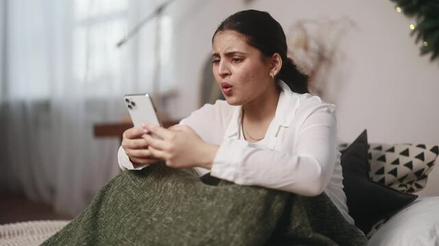 Sad young indian woman sitting on bed at home get notification and feeling upset reading bad negative news message on smartphone indoors Disappointment Fail Unemployment