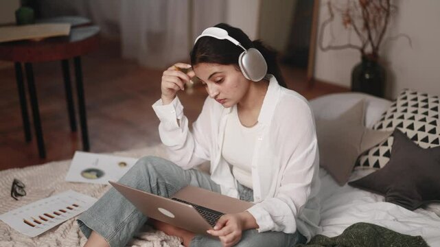 Boring unmotivated young indian woman student listening online lecture by laptop computer sitting on bed at home Tired girl with uninteresting lesson on remote distance education indoors