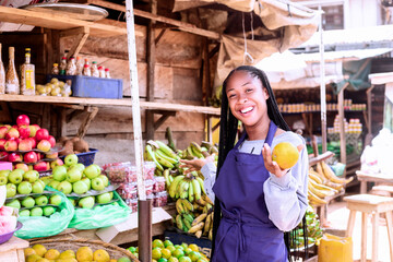 A cheerful vendor stands at her fruit stall, displaying a fresh orange, surrounded by vibrant produce.