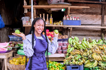 Cheerful girl wearing an apron showcasing fresh produce, including bananas and apples, in a lively african market setting.
