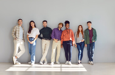 Obraz premium Group portrait of smiling young people standing near wall in looking into camera. Diverse group of happy multiracial and multinational students in casual clothes posing in brightly lit studio.