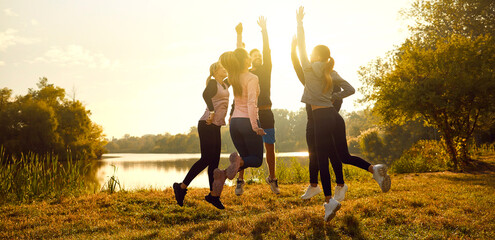 Group of sporty people in sportswear standing in a circle on the lake shore jumping with hands up after workout in nature at sunset in the park. Outdoors training, fitness exercises concept. Banner.