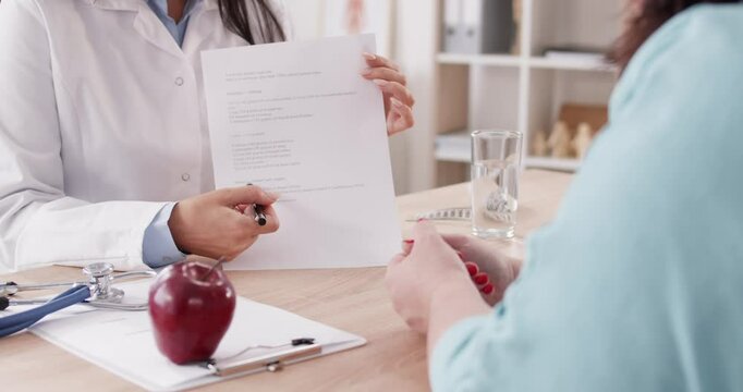 Close up of dietologist woman showing her fat woman patient diet plan and giving recommendations about healthy food and nutrition sitting on workplace in medical clinic. 4k video.