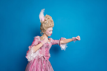Fototapeta premium Elegant woman in vintage baroque costume poses dramatically against a vibrant blue backdrop