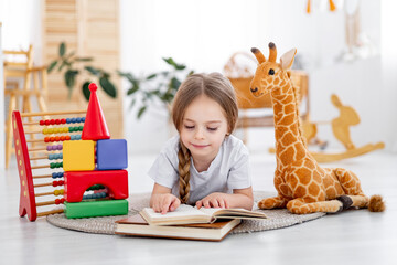 a little girl among the toys in the children's bright room at home or in kindergarten is reading a book or learning to read, a smiling child, the lifestyle of a preschooler child
