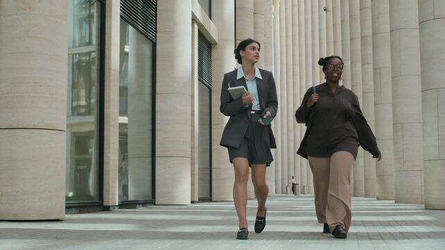 Low angle full shot of two women in formal outfits talking and strolling along paved walkway lined with tall columns of modern building
