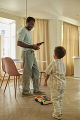 Vertical shot of little African American boy pointing at building blocks on toy cart paying dads attention while having fun at home together on weekend