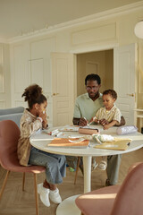Vertical shot of loving African American father with little son sitting on knees spending time together with kids drawing colorful pictures at table in cozy living room, copy space