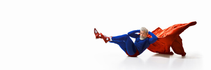 Grey-haired young man with beard, in superhero costume and reed cape, doing bicycle crunches, press exercises against white studio background. Concept of sport, healthy lifestyle, workout. Banner, ad © master1305