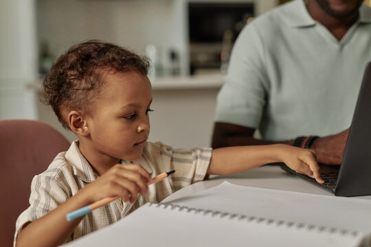 Medium shot of little Black boy pressing button of laptop keyboard curious about how to use computer while distracting father from work at home, copy space