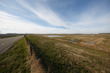 Wide landscape featuring a rural road beside a large field and a small water body under a clear sky during daylight