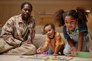 Excited African American boy throwing dice playing board game with dad and sister having fun together at home on weekend