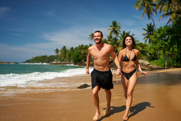 Joyful barefoot couple runs on sandy beach by ocean. Man and woman in swimwear enjoy tropical coast run. Fun, sunbathing, fitness beachside. Cheerful duo exercise, laugh near sea on sunny day.
