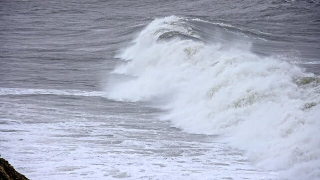 The ocean is rough and choppy, with waves crashing against the shore. The water is a deep blue color, and the sky is overcast. Scene is one of power and unpredictability