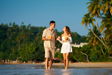 Couple stroll barefoot on sandy beach, holding coconuts, sipping drinks, enjoying sunset. Casual date, tropical vacation vibe. Smiles, flirty conversation, ocean backdrop, intimate walk together.