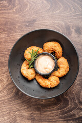 A black bowl featuring crispy fried shrimp arranged around a small cup of dipping sauce, garnished with a sprig of rosemary, set on a wooden table. Perfect for seafood lovers.