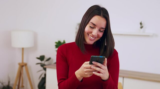 Smiling woman relaxing at home using cell phone device. Beautiful millennial female chatting on mobile phone application. Social media and technology concept.
