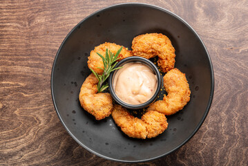 A black bowl featuring crispy fried shrimp arranged around a small cup of dipping sauce, garnished with a sprig of rosemary, set on a wooden table. Perfect for seafood lovers.