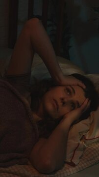 Vertical video. Young woman suffering from insomnia and headache lying in bed at night.