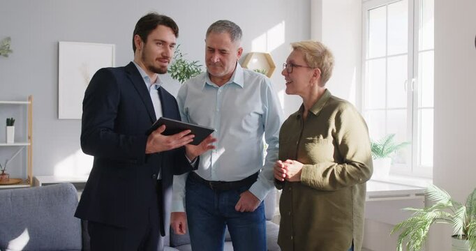 Young male real estate agent giving a tour about the house or apartment to a nice retired senior family couple investors. Real estate business concept 