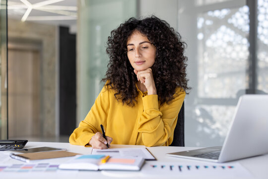 Serious focused woman doing paperwork inside office. Businesswoman filling in data, writing down paper reports. Financier thinking in office at workplace with laptop.