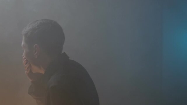 Man covering his mouth while suffocating in a smoke-filled room caused by a fire, coughing and struggling to breathe, unaware of the exit in the hazardous environment.