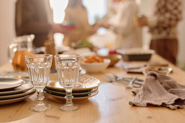 Medium close up of pair of polished glasses, stocked set of plates and homemade food in blurred background o wooden table