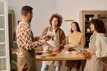 Biracial friends chatting all together while serving table, women putting baking tray and jar with juice on table while men polishing silverware