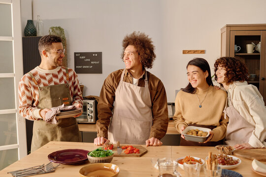 Wide shot of biracial company of friends laughing and smiling while serving wooden table with homemade food to celebrate house-warming party